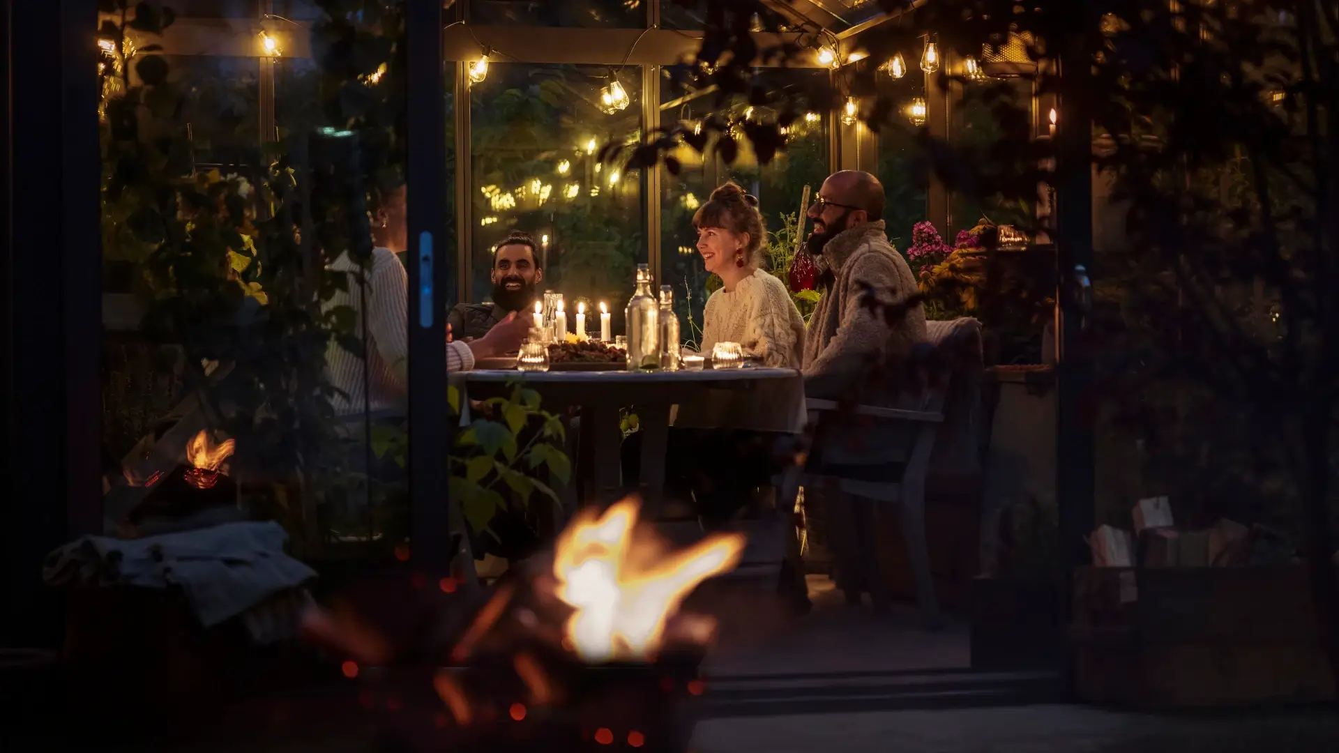 A group of people laughing and sharing a meal at a warmly lit dinner table inside a glass-enclosed space.